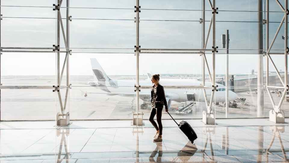 Reisende mit Rollkoffer geht durch den Flughafen, während draußen ein Flugzeug zu sehen ist.